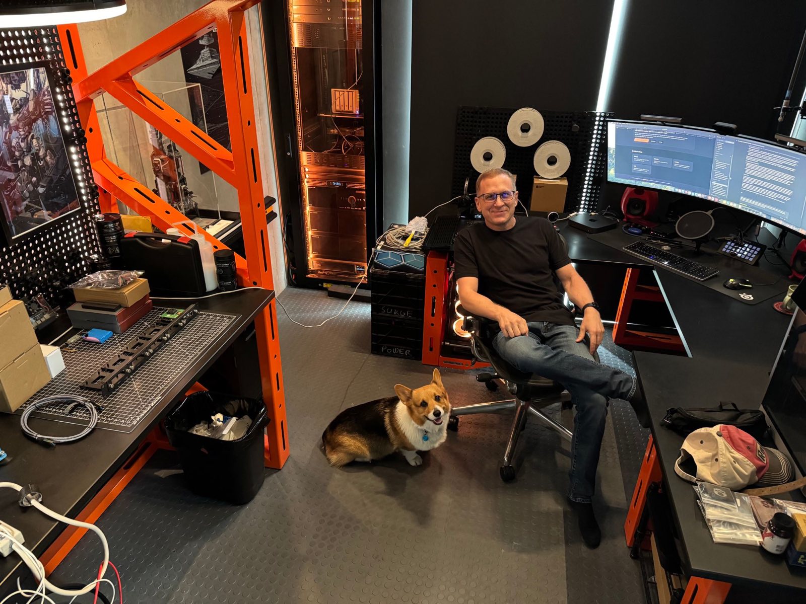 Bryce in the geekdojo workshop — workbench and electronics lab on the left, server rack in the background, a Pembroke Welsh Corgi sitting by the desk